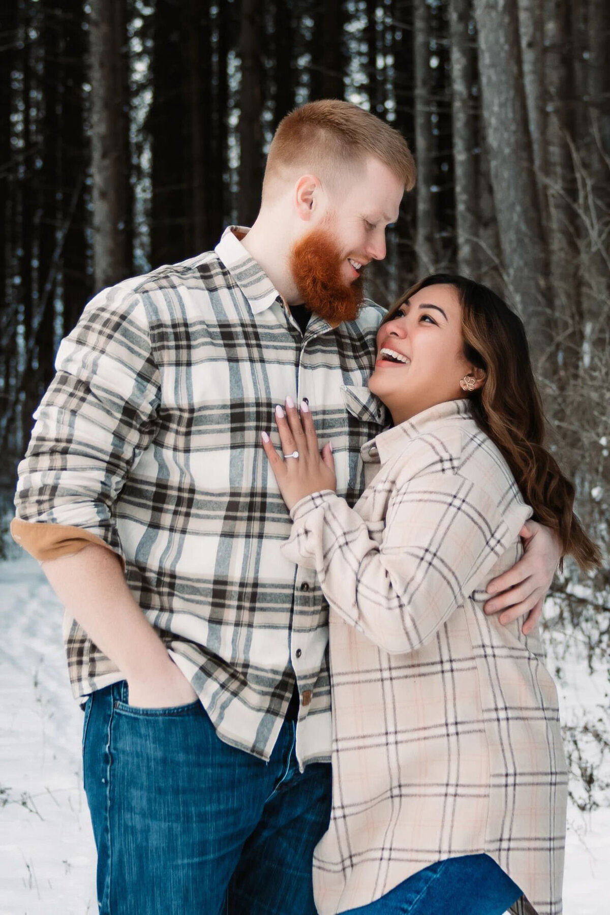 mand and woman snuggled up in the snowy forest while wearing plaid and laughing