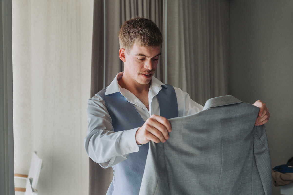 Groom smiles as he holds up his wedding suit jacket while getting ready at Lesner Inn in Virginia Beach, Virginia.