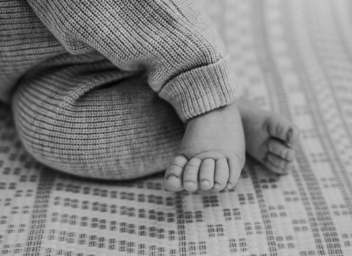 Newborn Photographer, a baby wears a knit onesie and lays on bed legs crossed