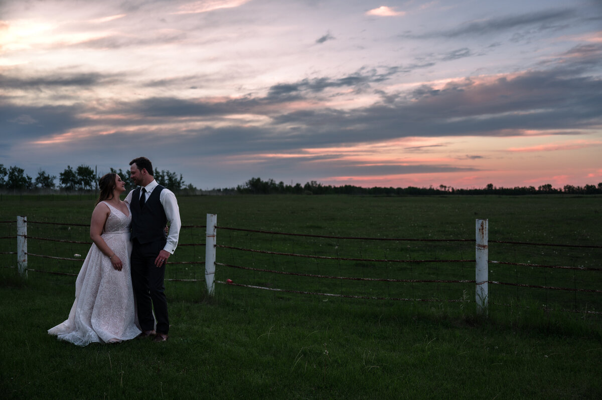bride and groom standing in a field with pink and orange sunset in the background