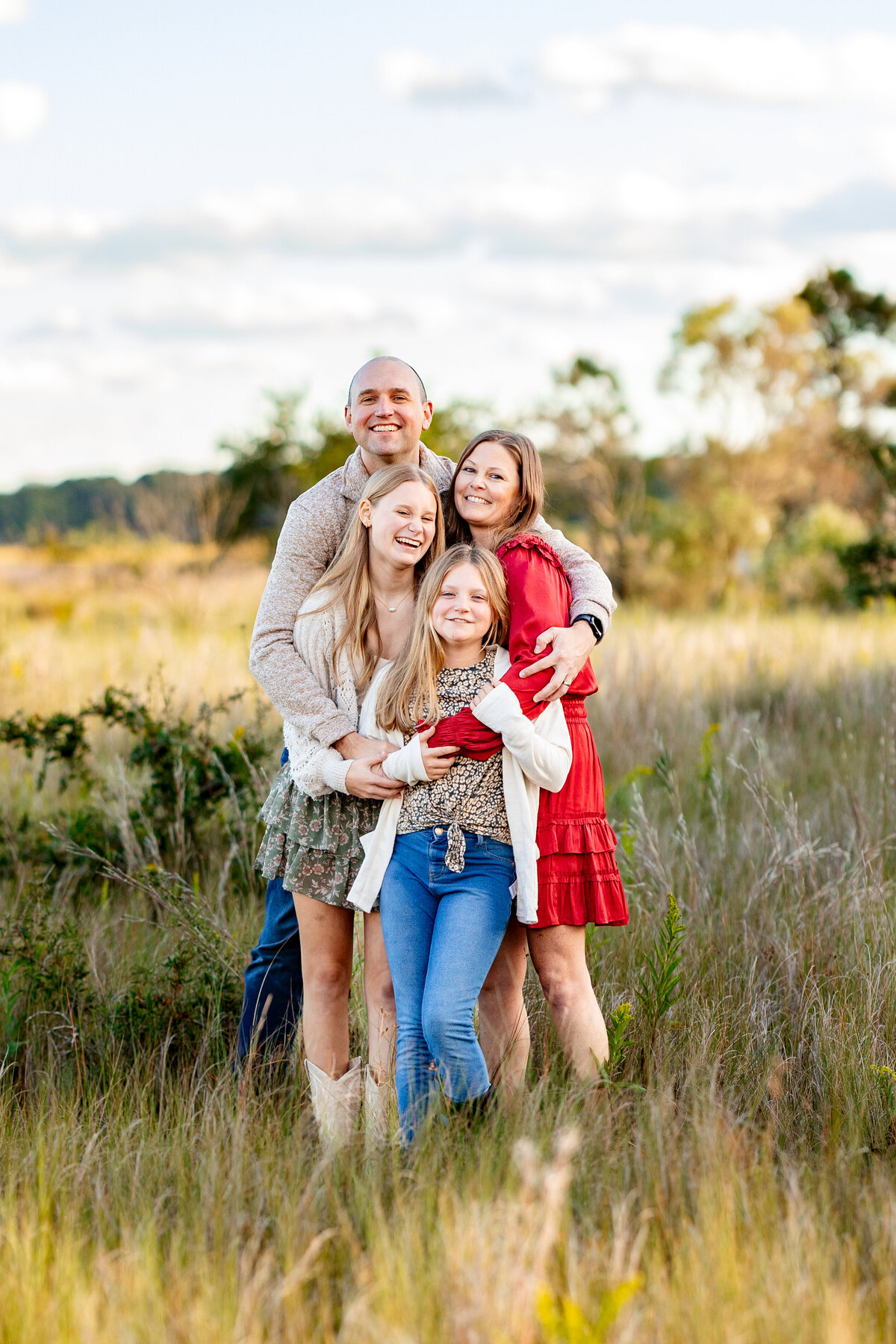 a vibrant family of four dressed in matching outfits cuddles together in the golden light of a wide grassy field laughing together during a family photography session at the Brock Nature Center in Virginia Beach, Virginia.  