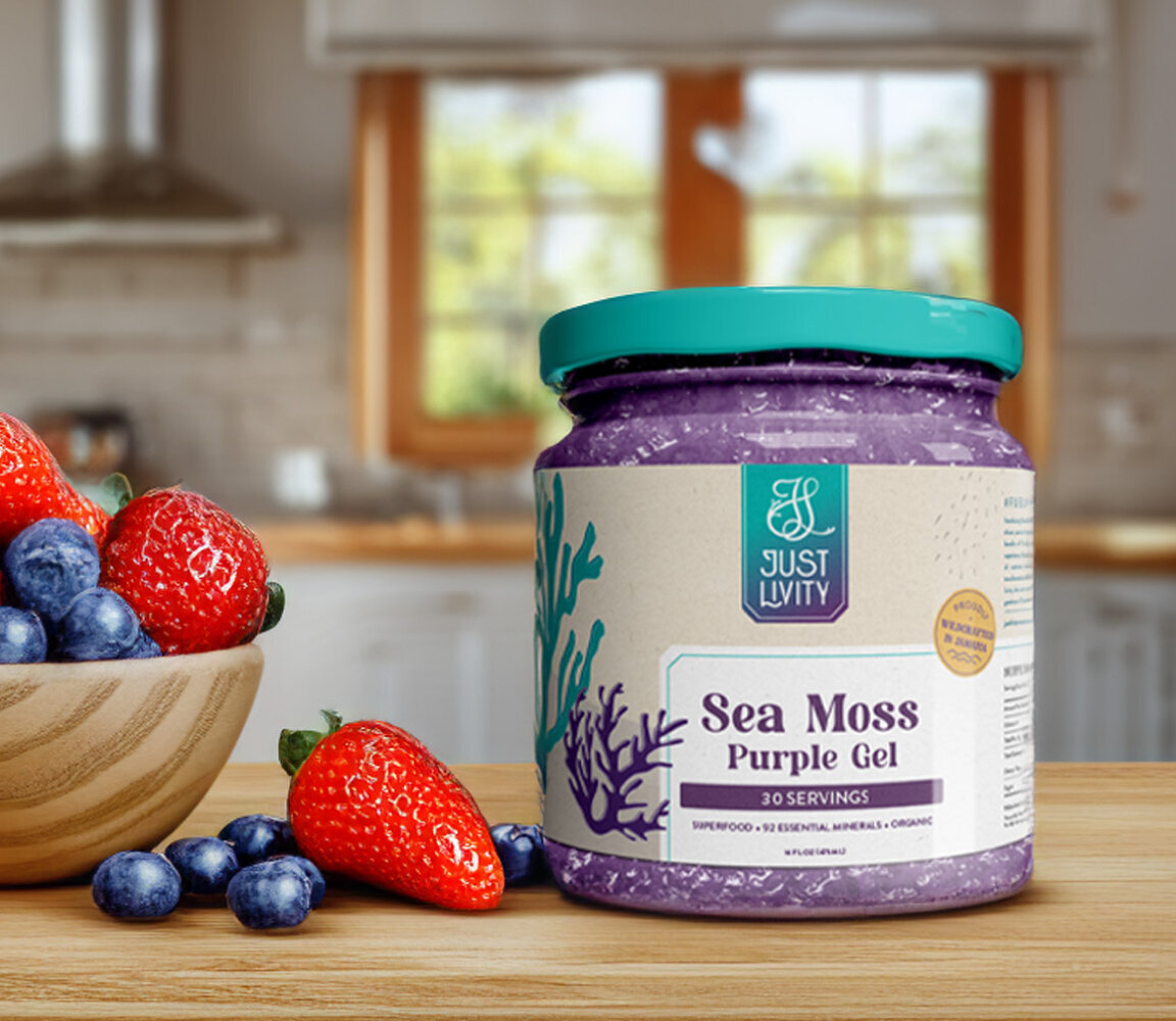 Mason jar with purple gel with packaging label for the brand Just Livity. The label showcases an underwater sea theme of sea moss. On the left side of the jar is a bowl of strawberries and blueberries. Both these items sit on a kitchen counter.