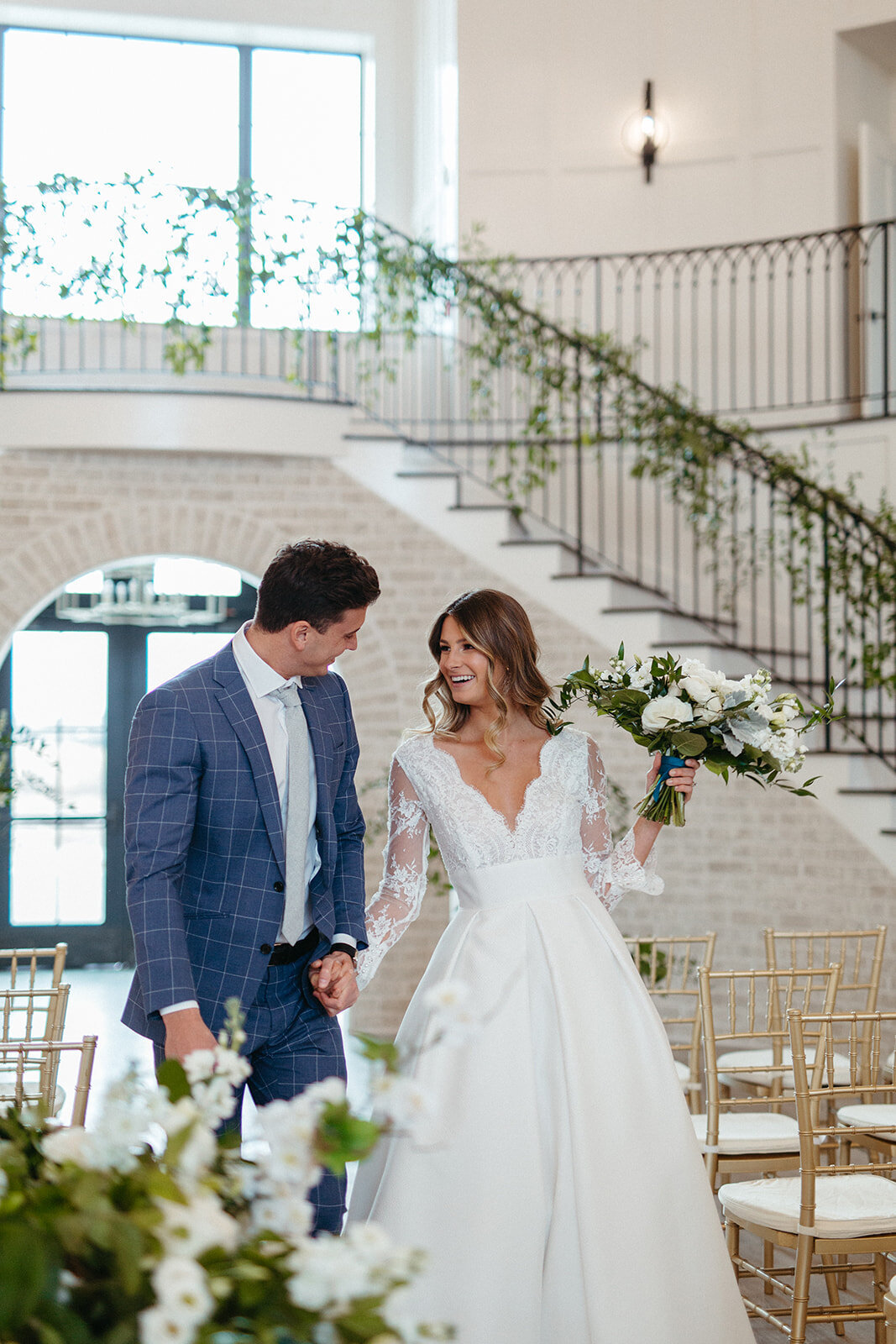 Bride and groom in  blue suit and white wedding gown walk down the aisle in a bright and airy room with gold chairs.