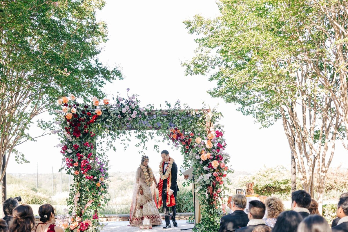 A bride and groom in traditional indian attire stand under a floral arch at an outdoor wedding ceremony, surrounded by guests.