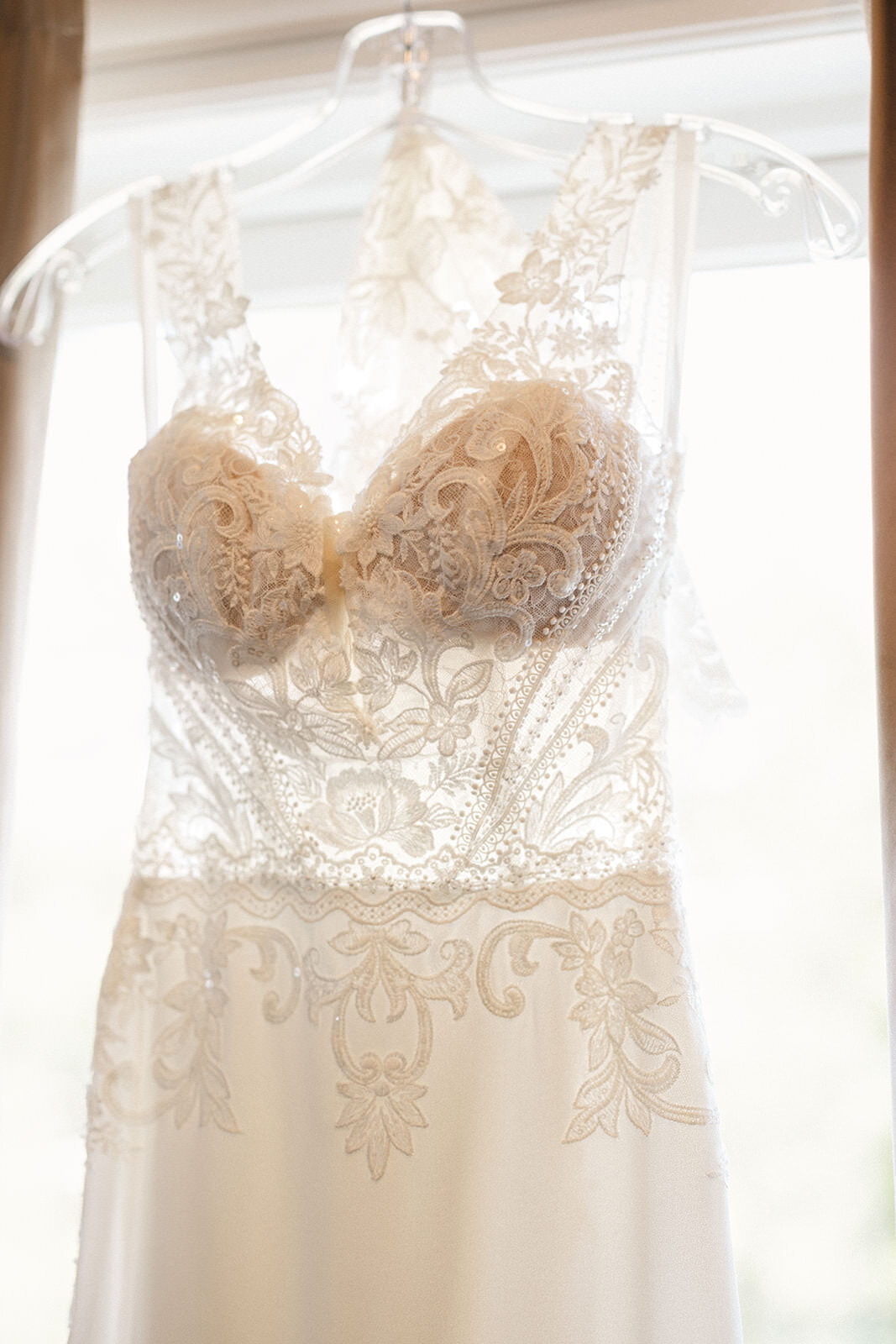 An elegant white wedding dress with lace detailing hanging in front of a window, radiating with natural light.