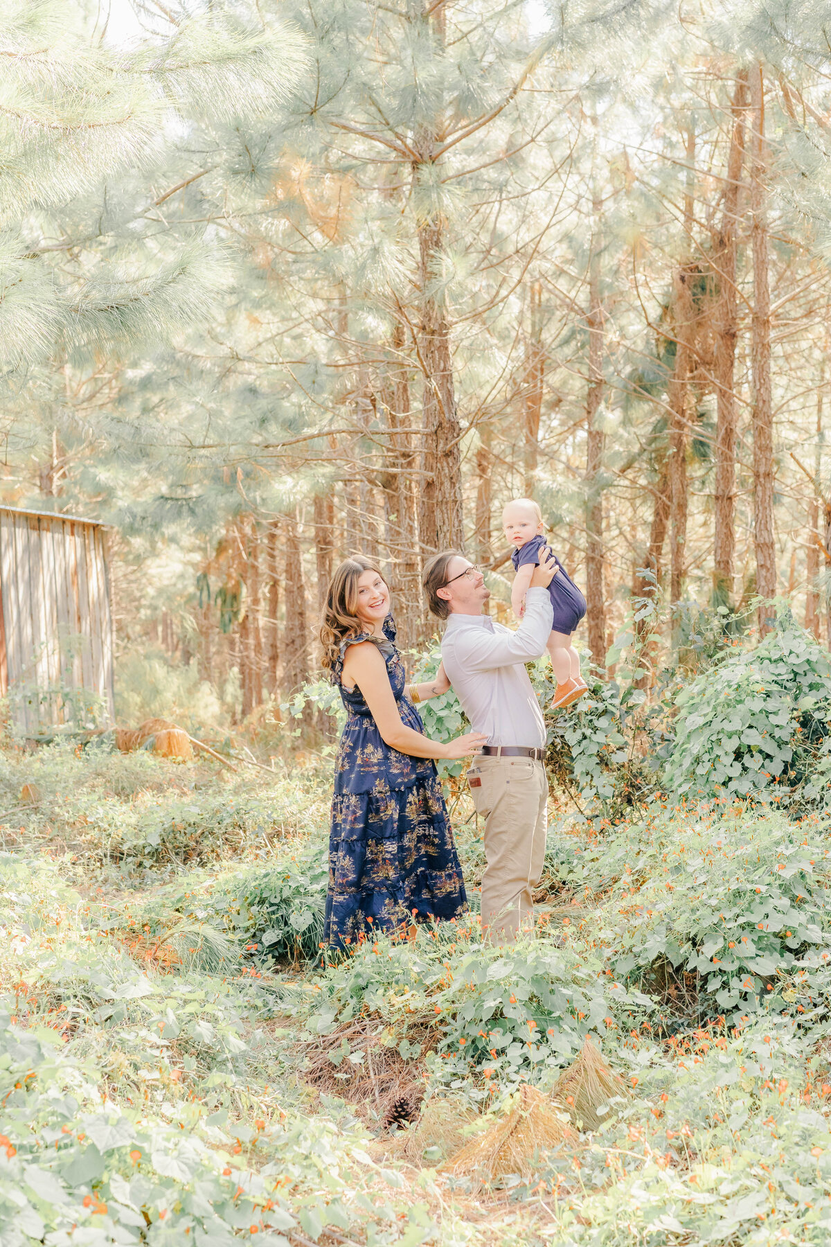 dad throwing baby in air while mom observes in wooded area