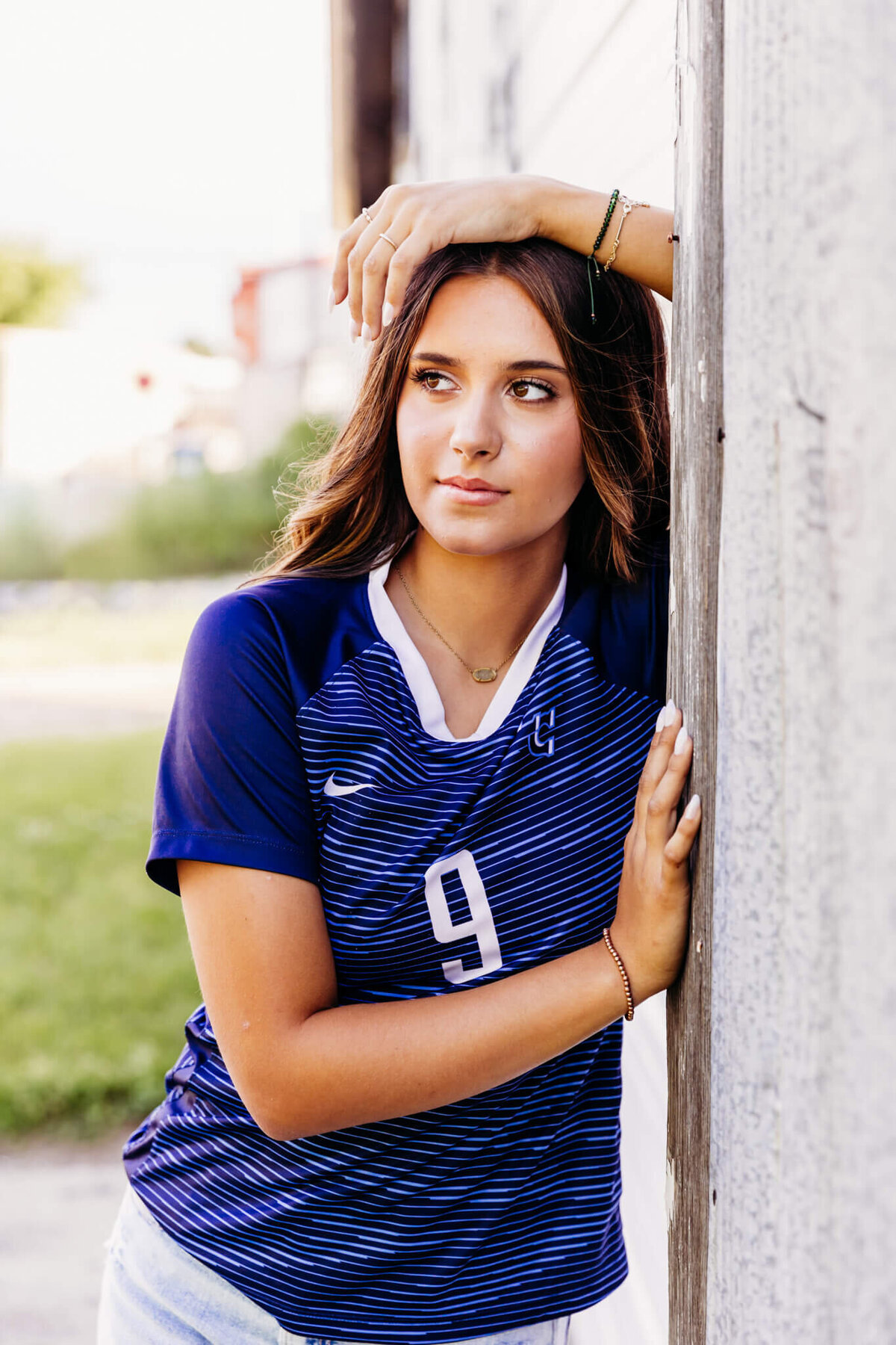 young teen girl in a blue soccer jersey leaning against a metal wall as she looks away captured by Ashley Kalbus Photography 