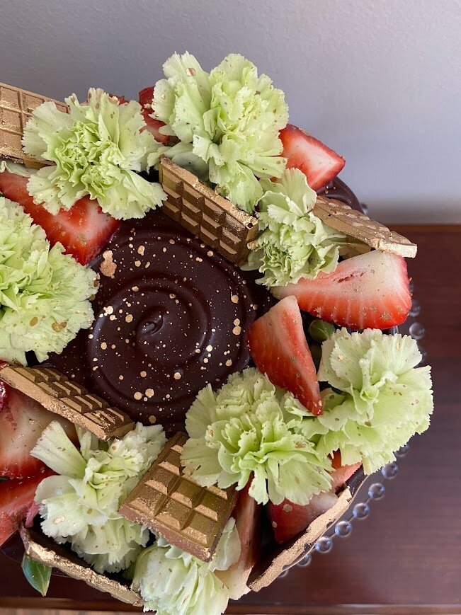 A close up shot of the top of a cake shows a ganache drip, with a wreath of chrysanthemum flowers, fresh strawberries, gold painted chocolate bars, and gold splatter.