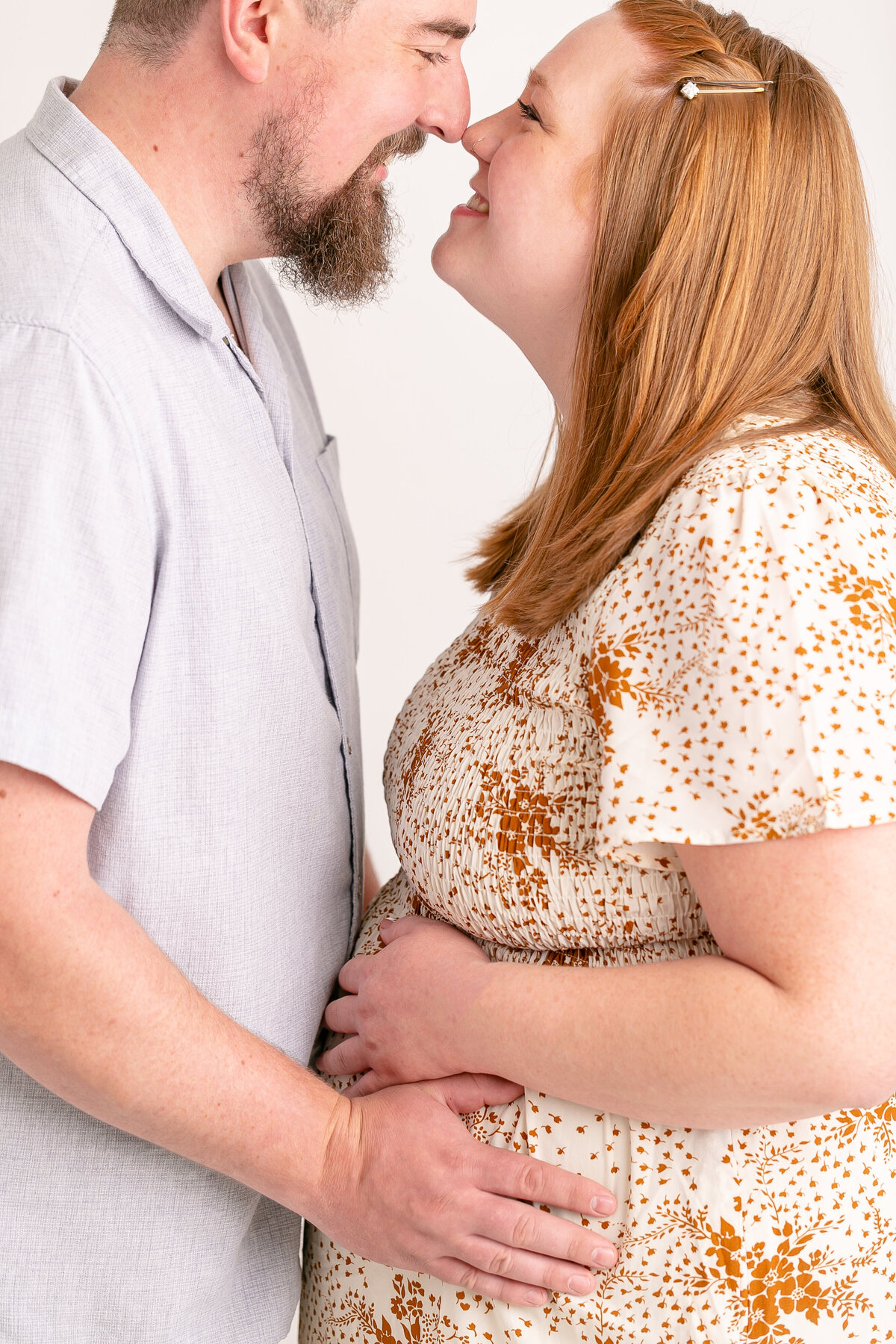 Man in baby blue shirt  standing nose to nose with pregnant wife. They are both holding pregnant belly together. Woman has red hair and is wearing a beige and burnt orange floral dress. Image taken in the studio of a Portland Maternity Photographer.