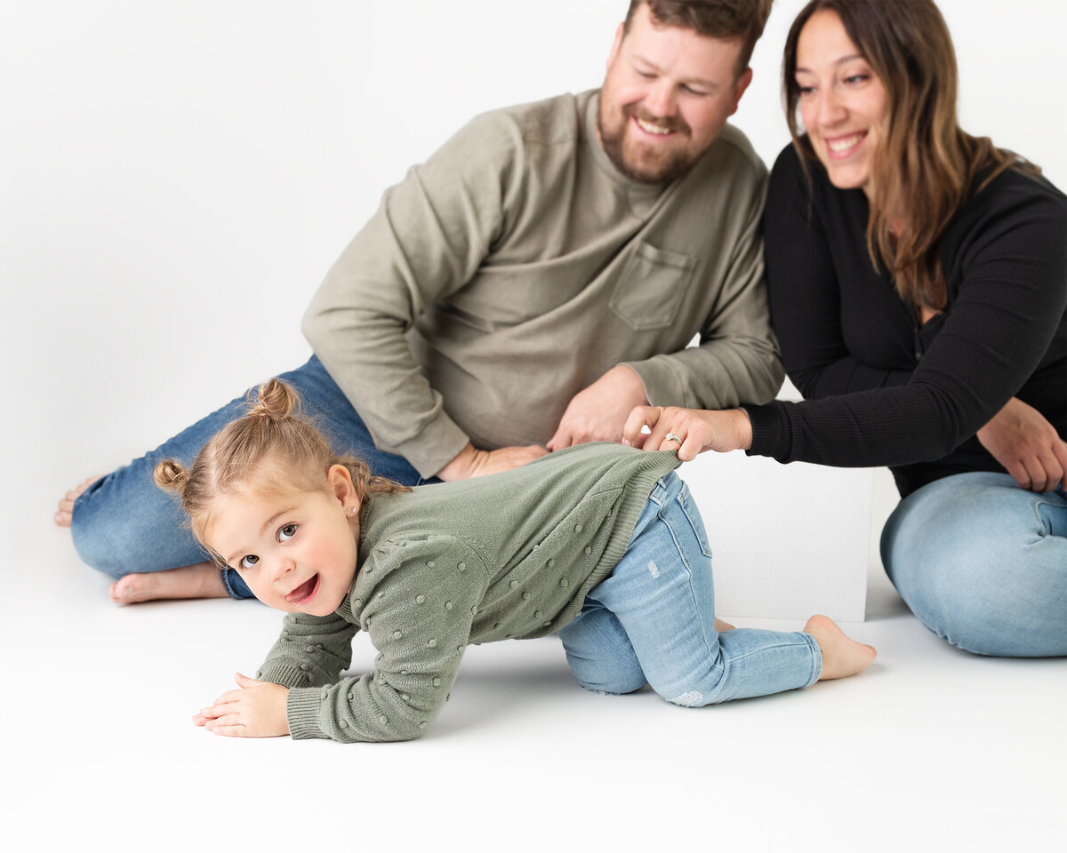 Toddler crawling and smiling toward the camera while parents laugh behind her in a white studio space in Paris, Ontario