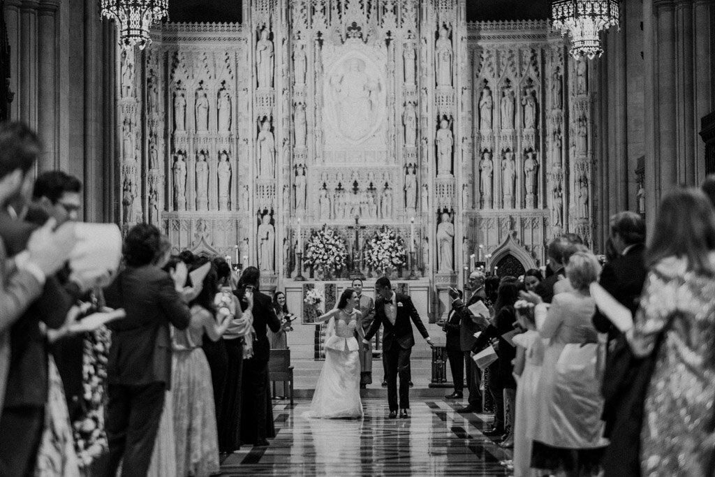 The photo was taken at the Mellon Auditorium in Washington D.C., one of the best luxury DC venues with European flair, by photographer Sarah Bradshaw. The wedding ceremony takes place in a grand, ornate church. The bride and groom, dressed in formal attire, walk hand in hand down the aisle as guests cheer and take photos. The background features intricate carvings and large chandeliers, enhancing the elegant atmosphere of the occasion.