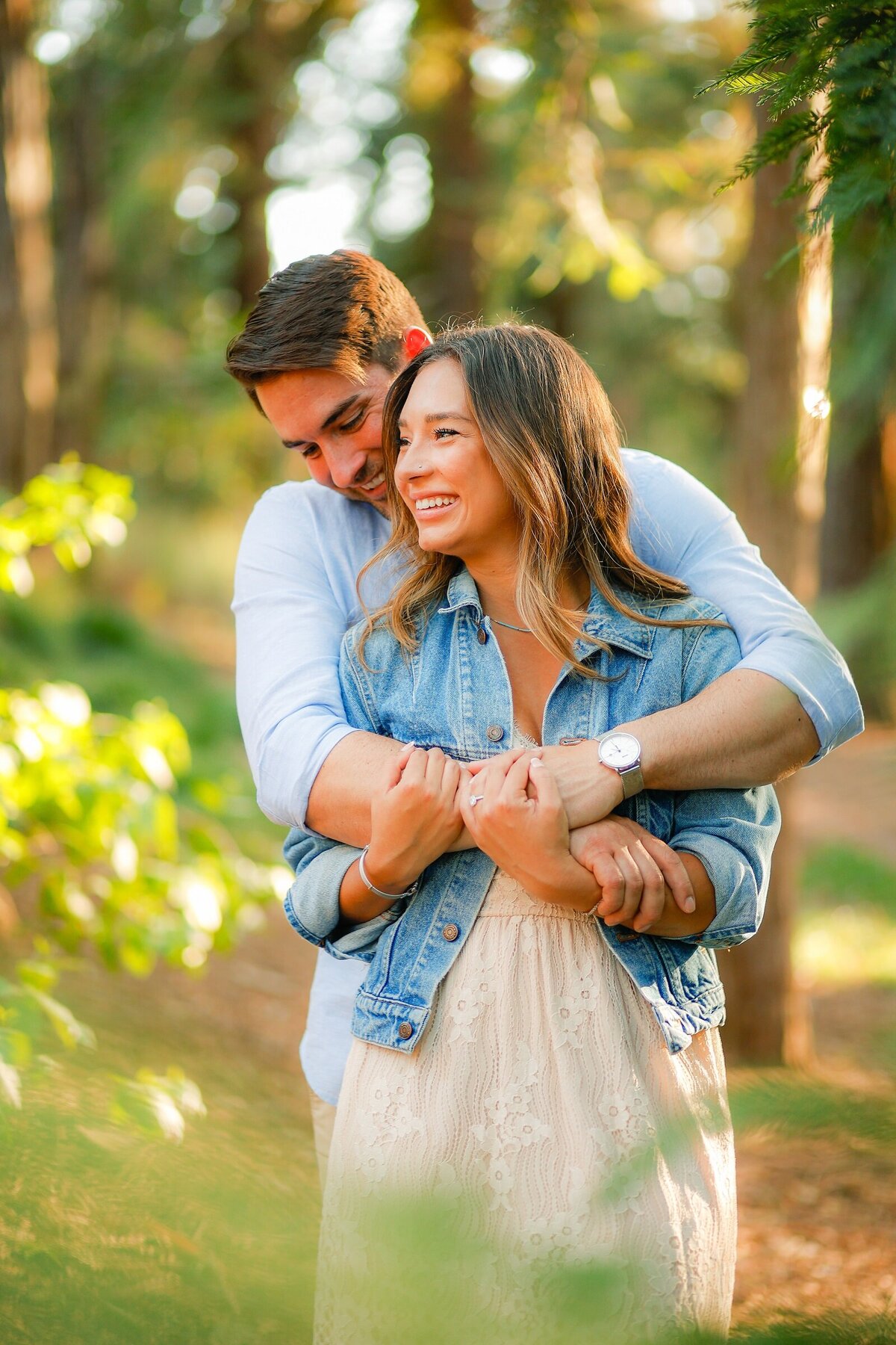 Photo of man hugging fiancé from  behind as she smiles and holds onto his arms. They are surrounded by trees at the UC Davis arboretum near sacramento, ca.