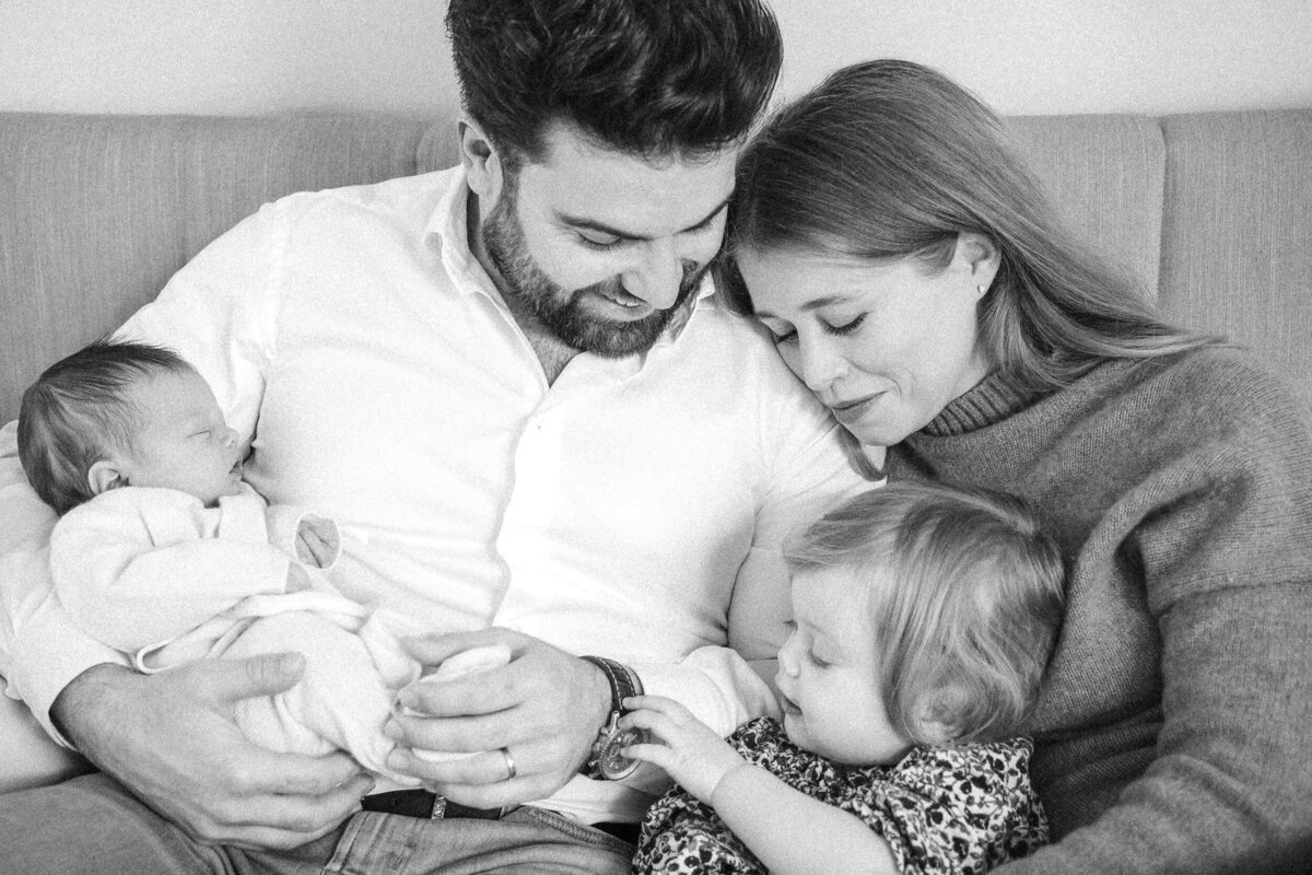 Father holding a newborn while mother rests her head on his shoulder and their toddler reaches for his watch.
