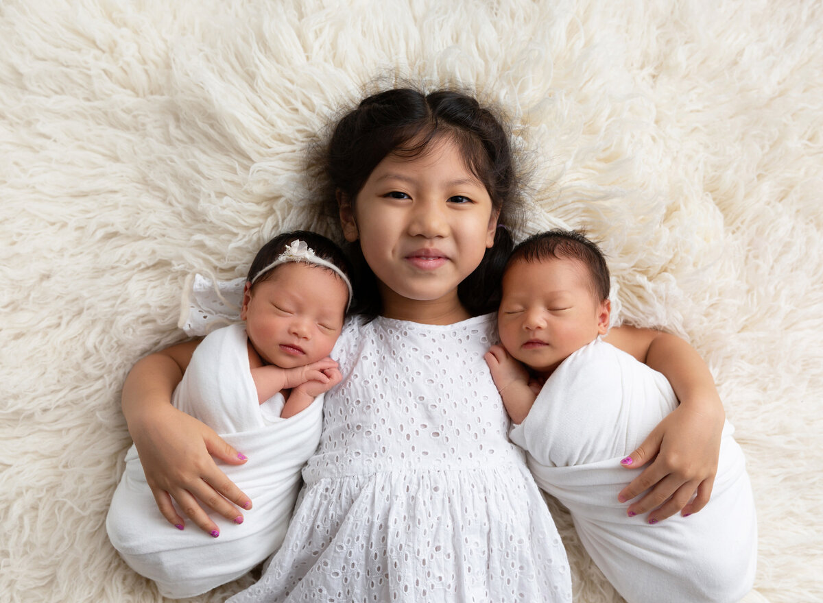 Aerial image captured during a newborn photoshoot.Big sister is holding her new bother and sister. The babies are resting their heads on sister's shoulder and her arms are wrapped around them.
