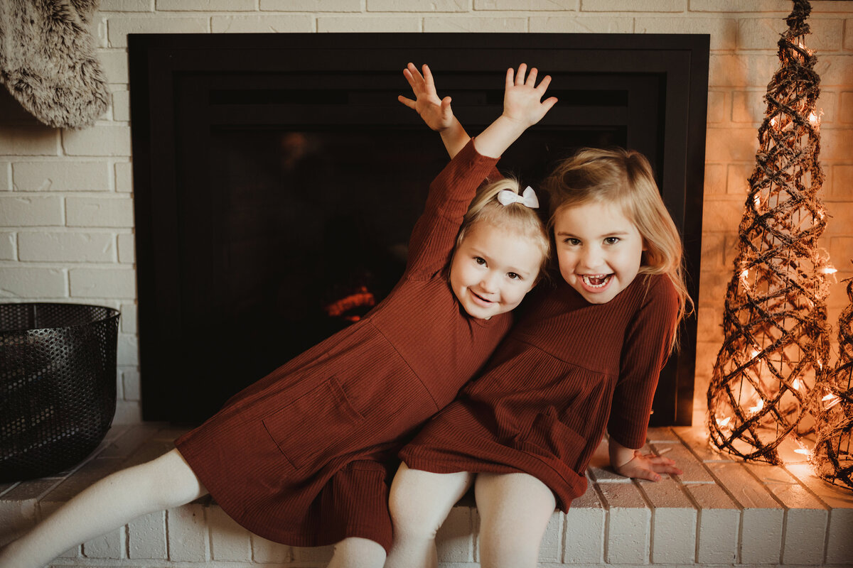 Two sisters sit in front of the fireplace, giggling and looking at the camera.