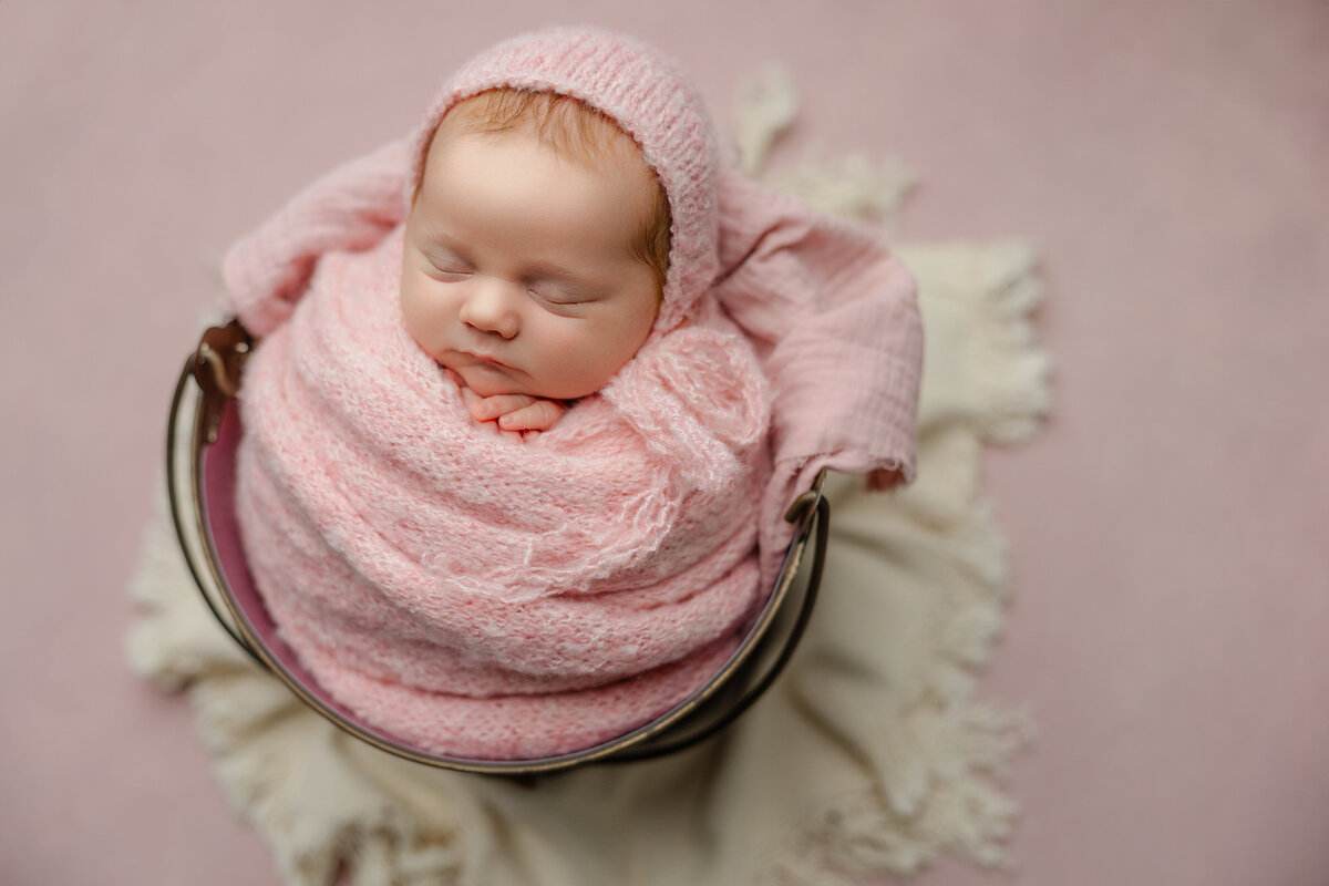 Studio newborn portrait featuring a swaddled baby girl in soft pink tones and classic bucket pose, styled on a pastel backdrop. Captured in the Inland Empire by Kaitlyn Dawn Photography.