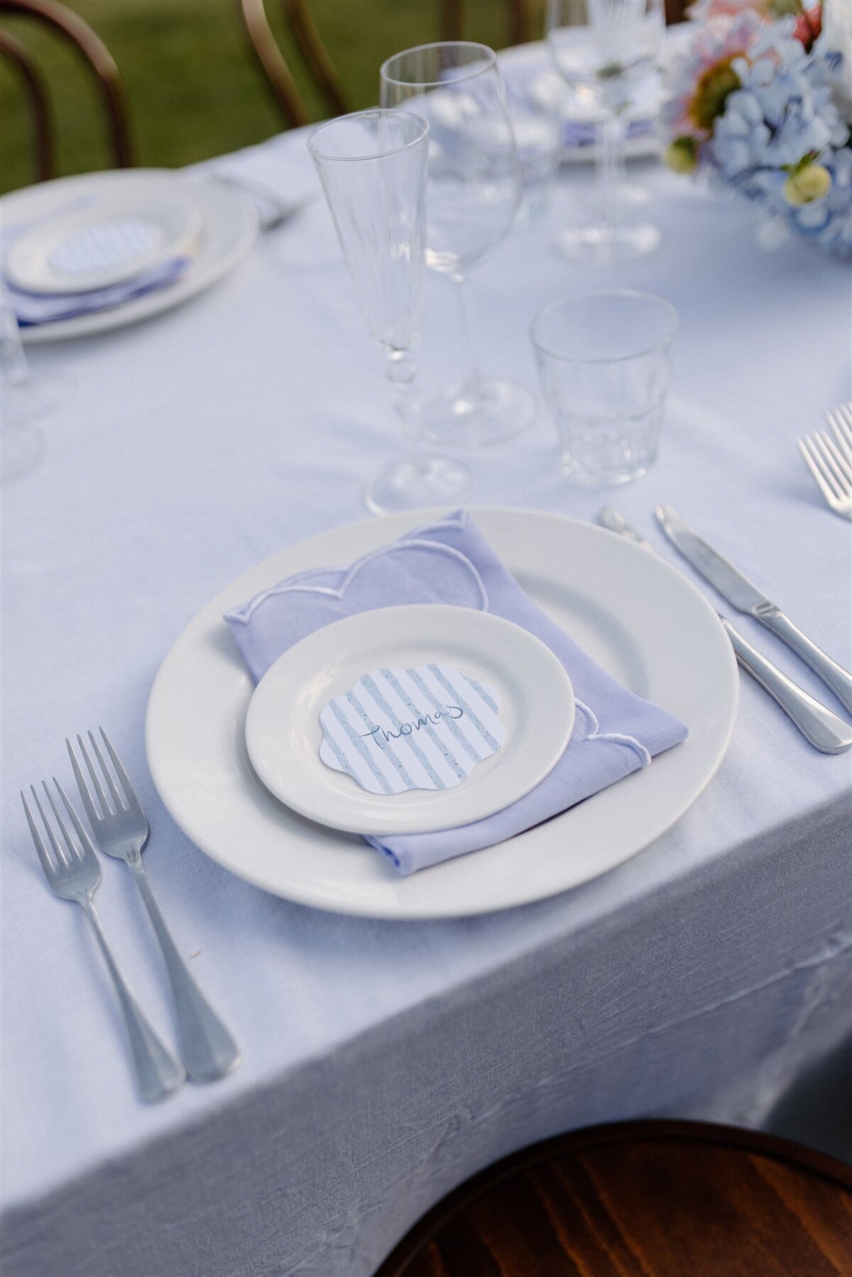 Place setting featuring blue striped name card, lilac napkin, and layered white plates on an elegant alfresco wedding table in Central Otago.
