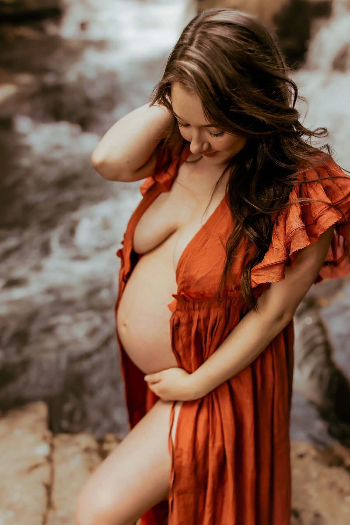 A pregnant woman in an orange dress stands beside a river, looking down and holding her belly.