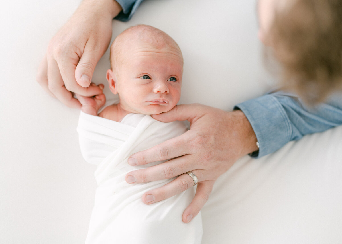 Baby swaddled in a white blanket looking up at his father who is holding his tiny fingers.