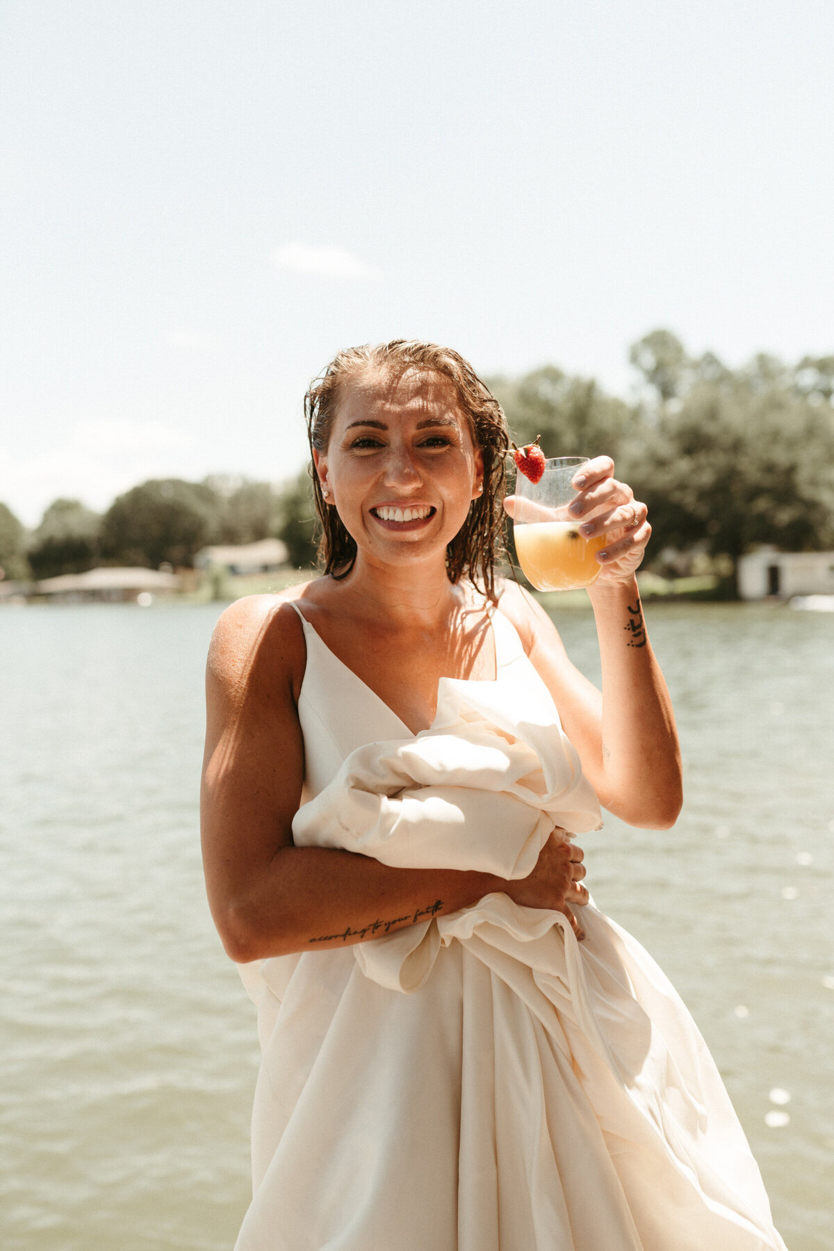 Bride with wet hair and wedding dress at a lake holding up her cocktail drink and smiling