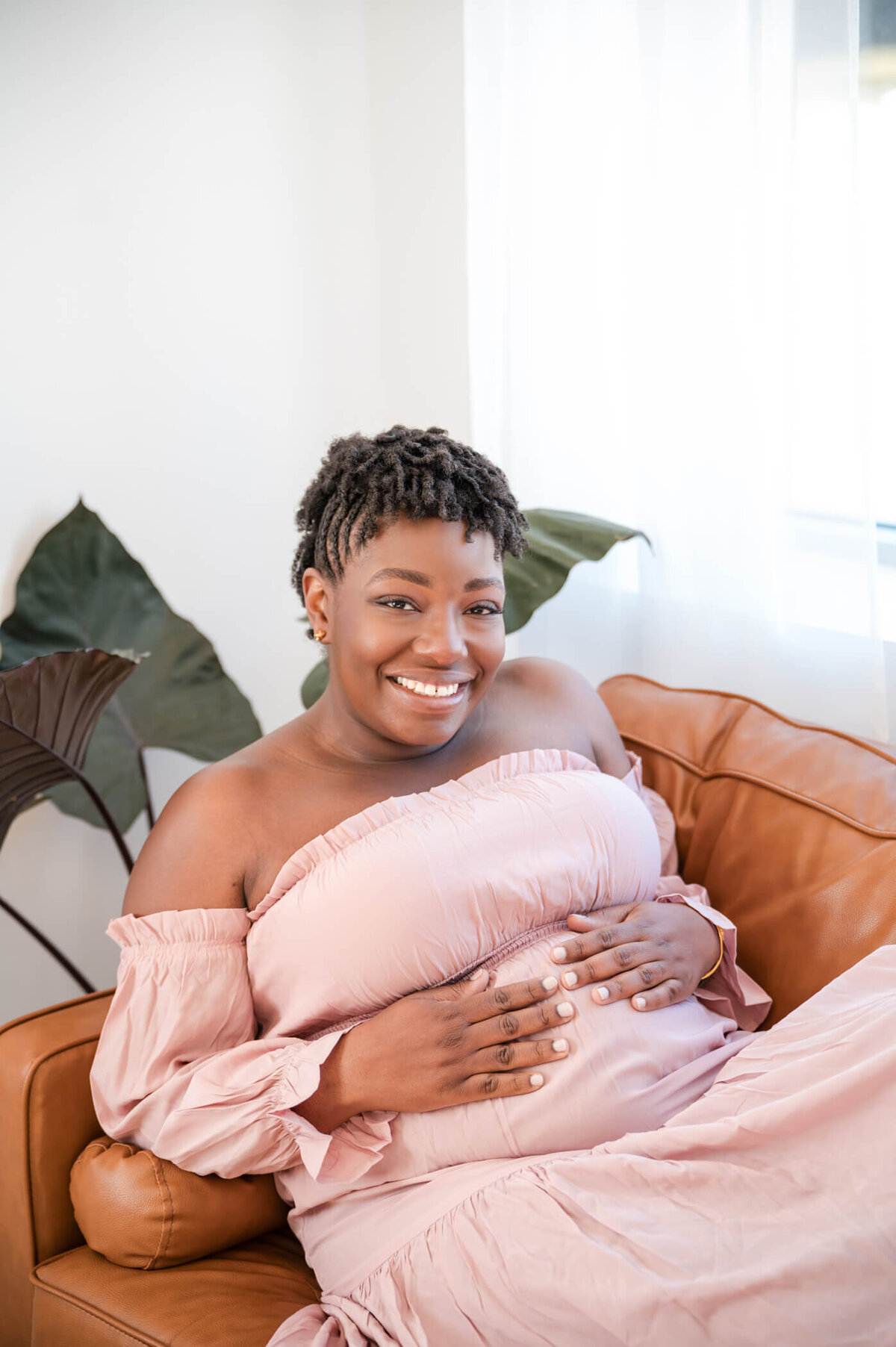 Expectant mother holding her belly while relaxing on a couch posing for maternity photos in a pink dress