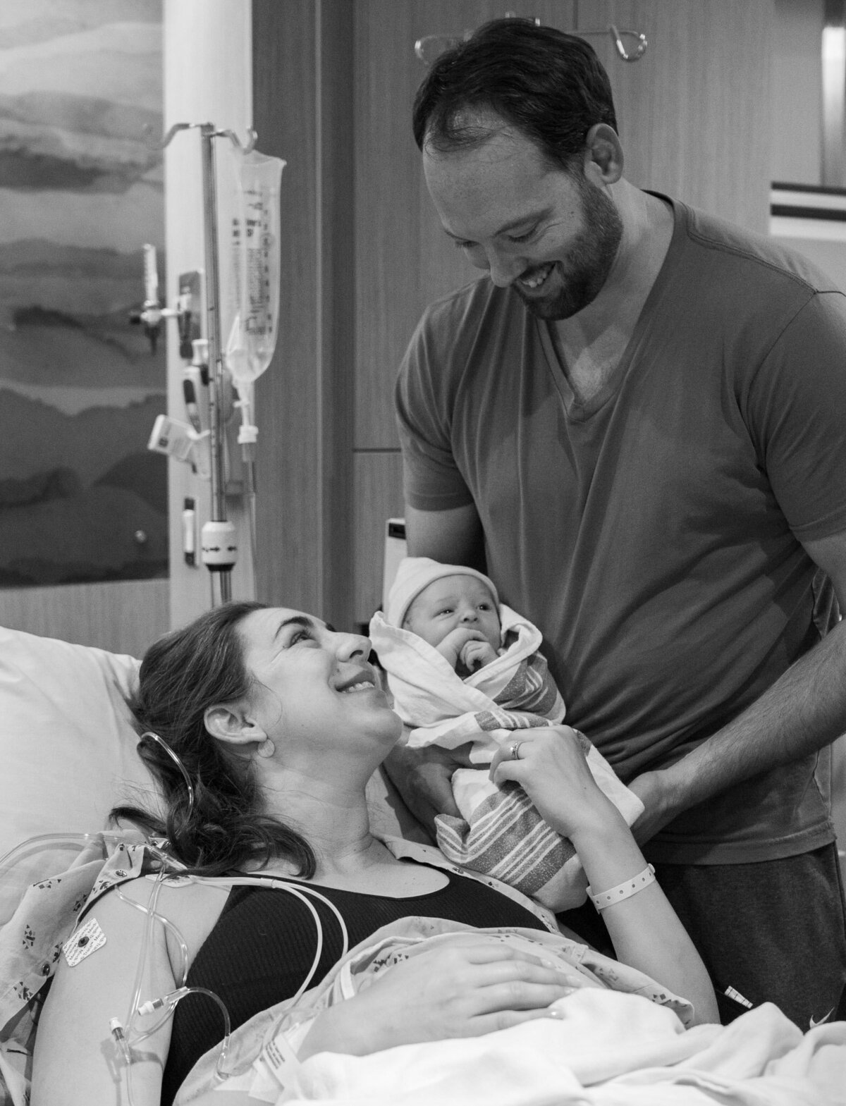 a man holds his new baby and smiles down at his wife in the hospital bed who has just given birth.