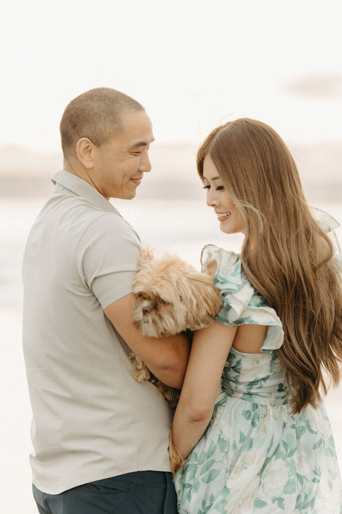 Couple standing together on the beach, man holding their small dog, as they smile at each other.