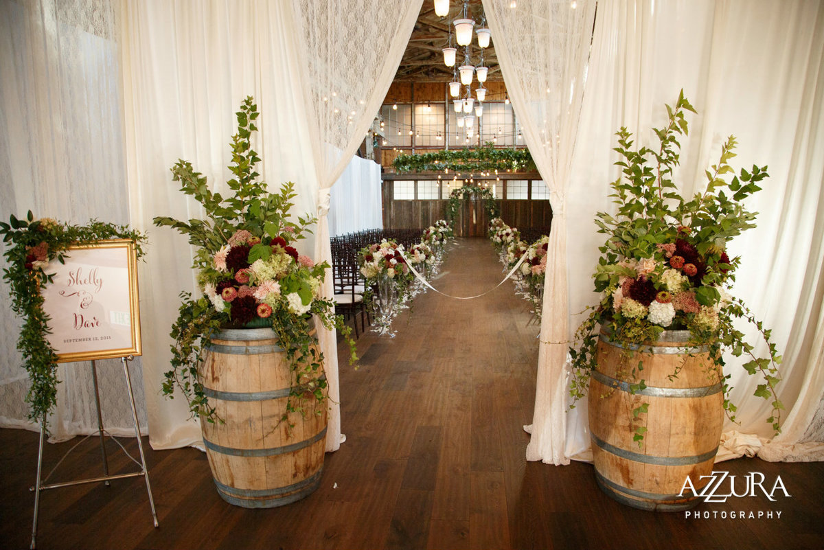 wedding ceremony entry decor with draping, barrels with large flower arrangements and welcome sign decorated in greenery vines