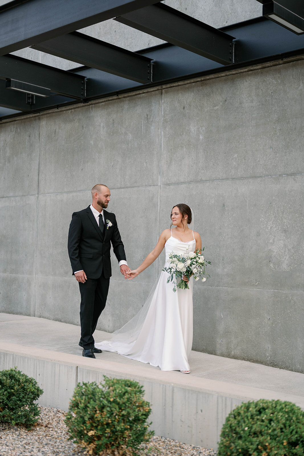 Bride and groom walking hand-in-hand along the brick wall entrance of Leona Road in Grand Rapids MI, captured during their stylish September wedding.