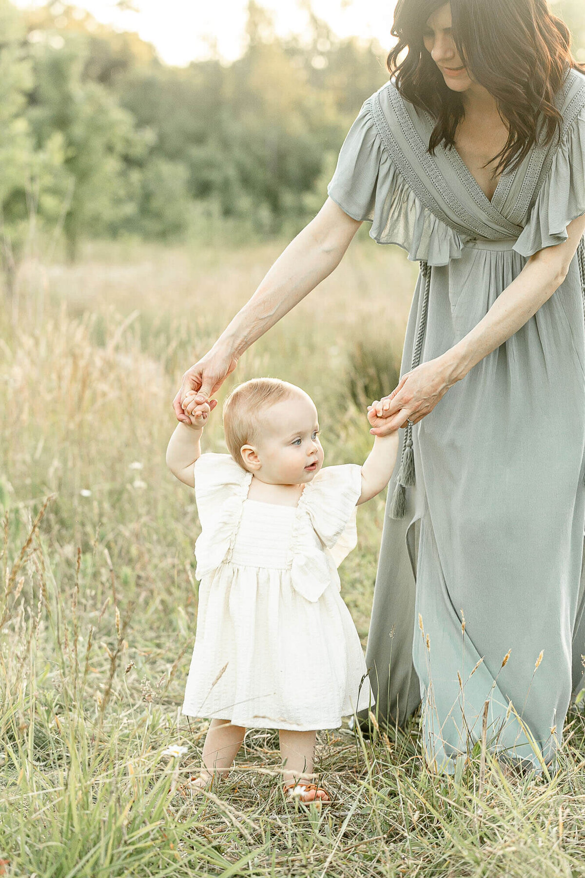Baby Girl just learning how to walk is dressed in a cute cream colored dress and brown sandals and is holding onto Mama's hands. Mama is wearing a sage green dress. They are standing out in a field of tall golden grasses during summertime at sunset.