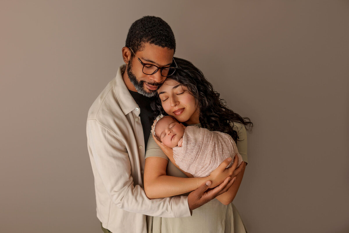 A loving couple holds their swaddled newborn baby, all three resting their heads closely together, radiating warmth and tenderness. The background is a neutral beige, enhancing the serene and intimate moment.