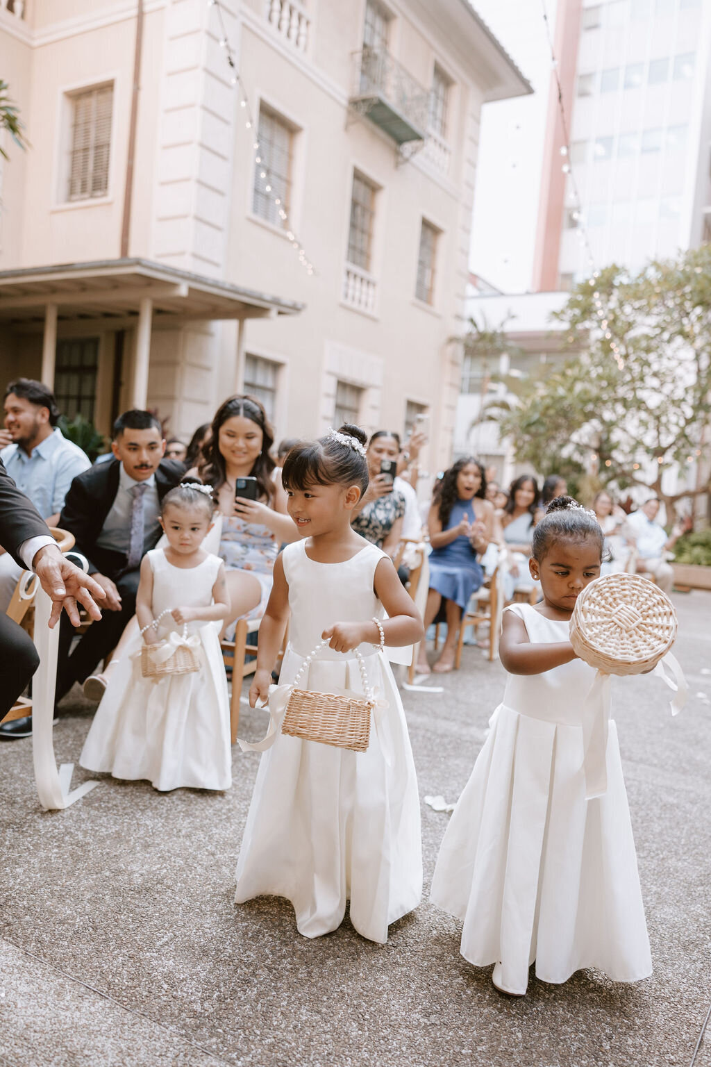Flower girls walking down the aisle in ivory dresses while mom takes phone photo in the background seated