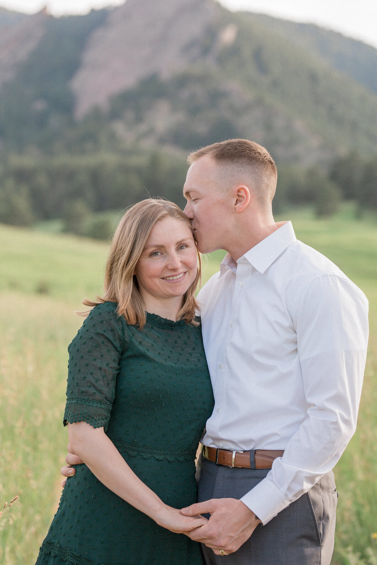 A man in a white button down shirt kisses his fiancee, dressed in green, on the temple