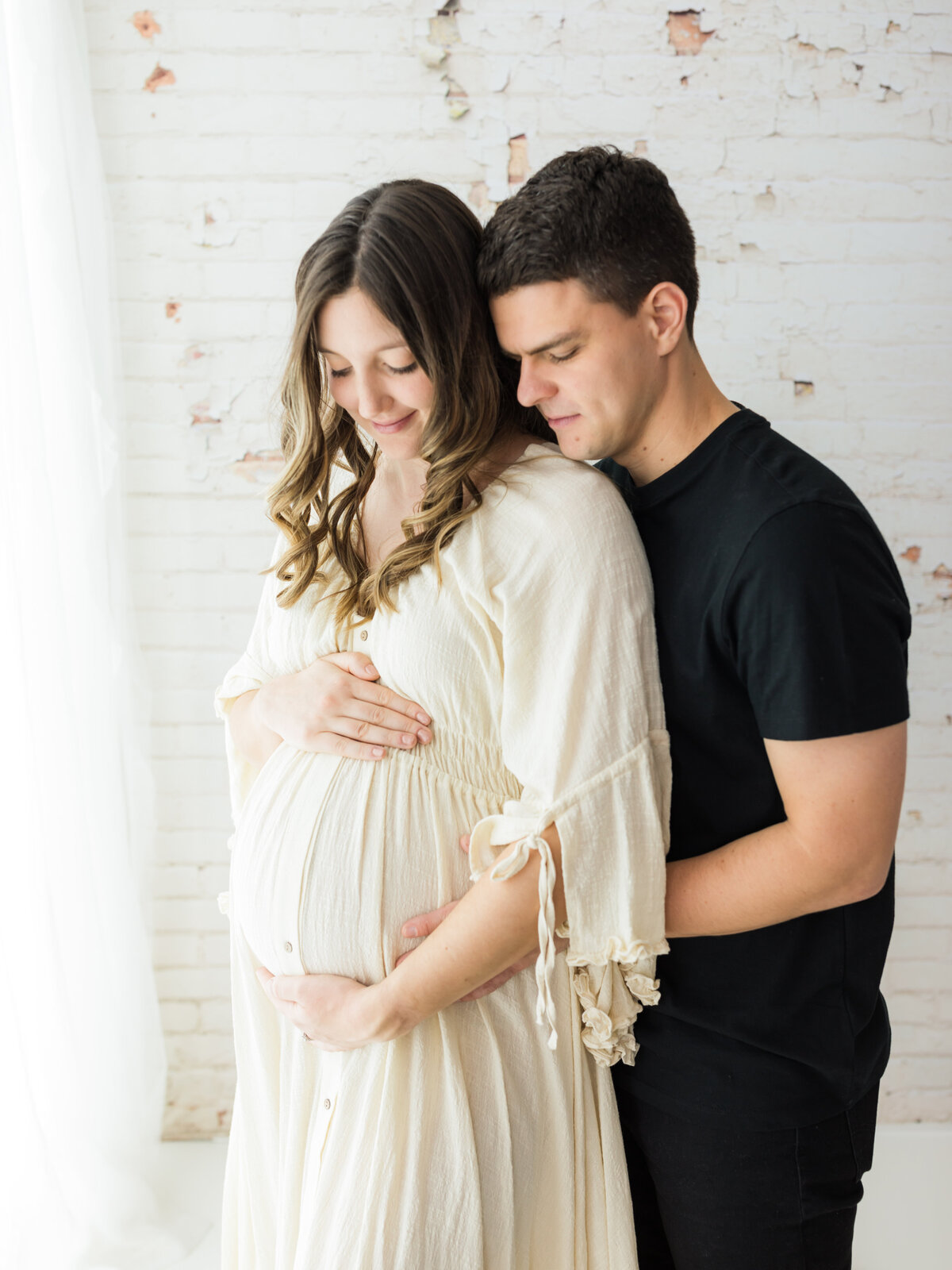 husband and pregnant  wife in cream maternity dress holding baby bump for maternity photoshoot