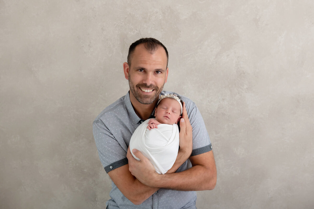 dad holding newborn baby girl wrapped in white for her newborn photos in Burlington