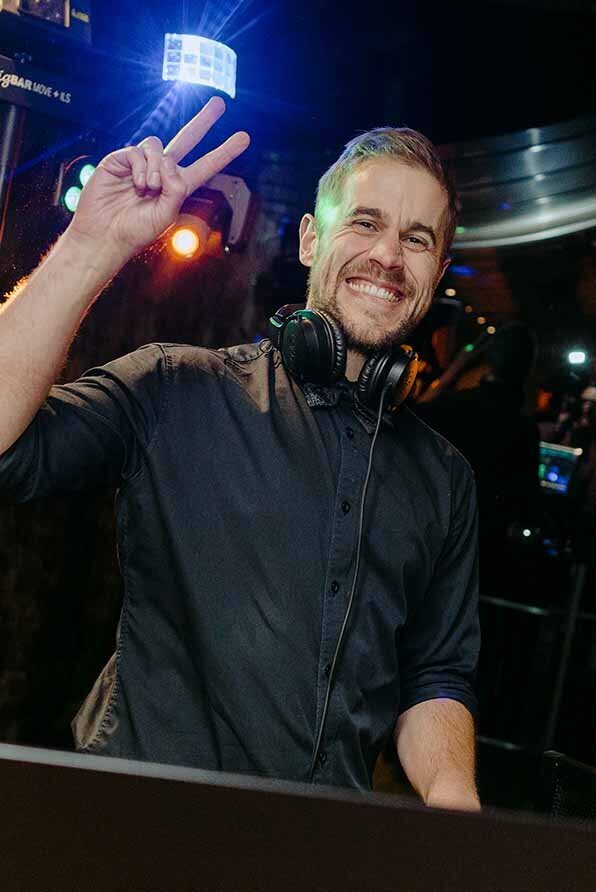 Male DJ smiling and signalling a peace sign behind the decks at a wedding reception with colourful flashing lights.