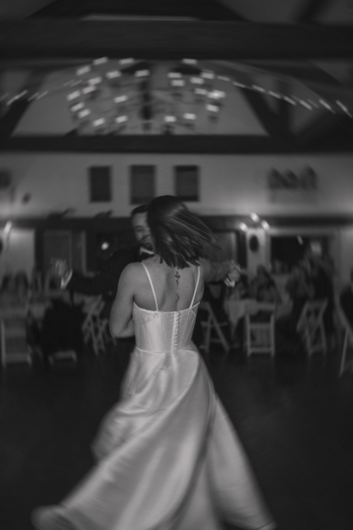 Bride and groom share a dynamic dance at their wedding reception, captured in a blurred motion that emphasizes the movement and energy. The bride's back tattoo is visible as her satin gown flows around her, while the groom holds her hand, both immersed in the joyous moment.