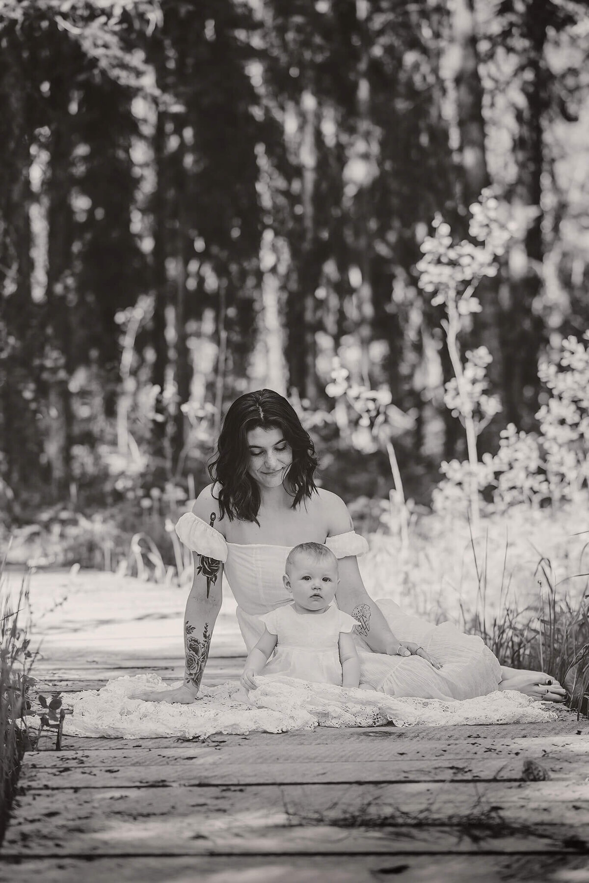 A black and white portrait of a mother and daughter sitting on a wooden bridge in the forest. Mom looks down at daughter while she smiles.  Taken by Brittany Danielle Photgraphy