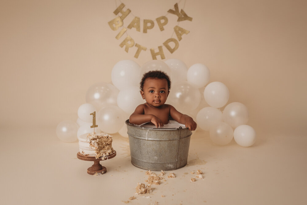 Photo shoot with Atlanta, Georgia cake smash photographer. Atlanta cake smash photography session at Marietta, Georgia portrait studio with one year baby in  small bubble bath tub with smash cake next to him and balloons and happy birthday sign in the backdrop.