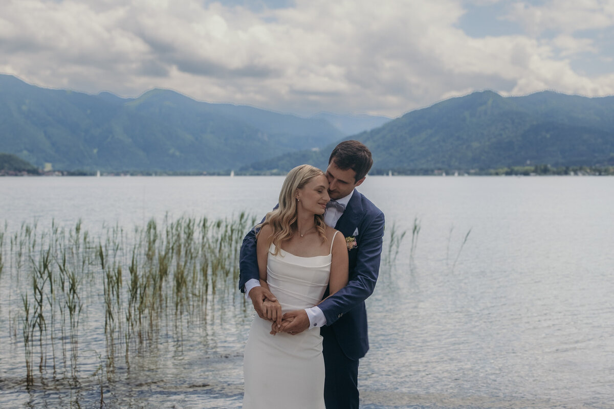 Bride and groom stand in a warm embrace in front of a big lake with mountains in the background.