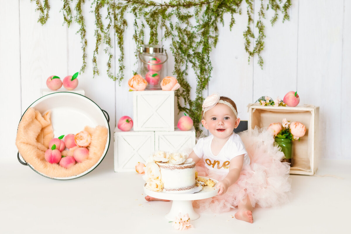 Cake Smash Photographer, a baby sits with a cake. There are pots  and boxes full with peaches behind her