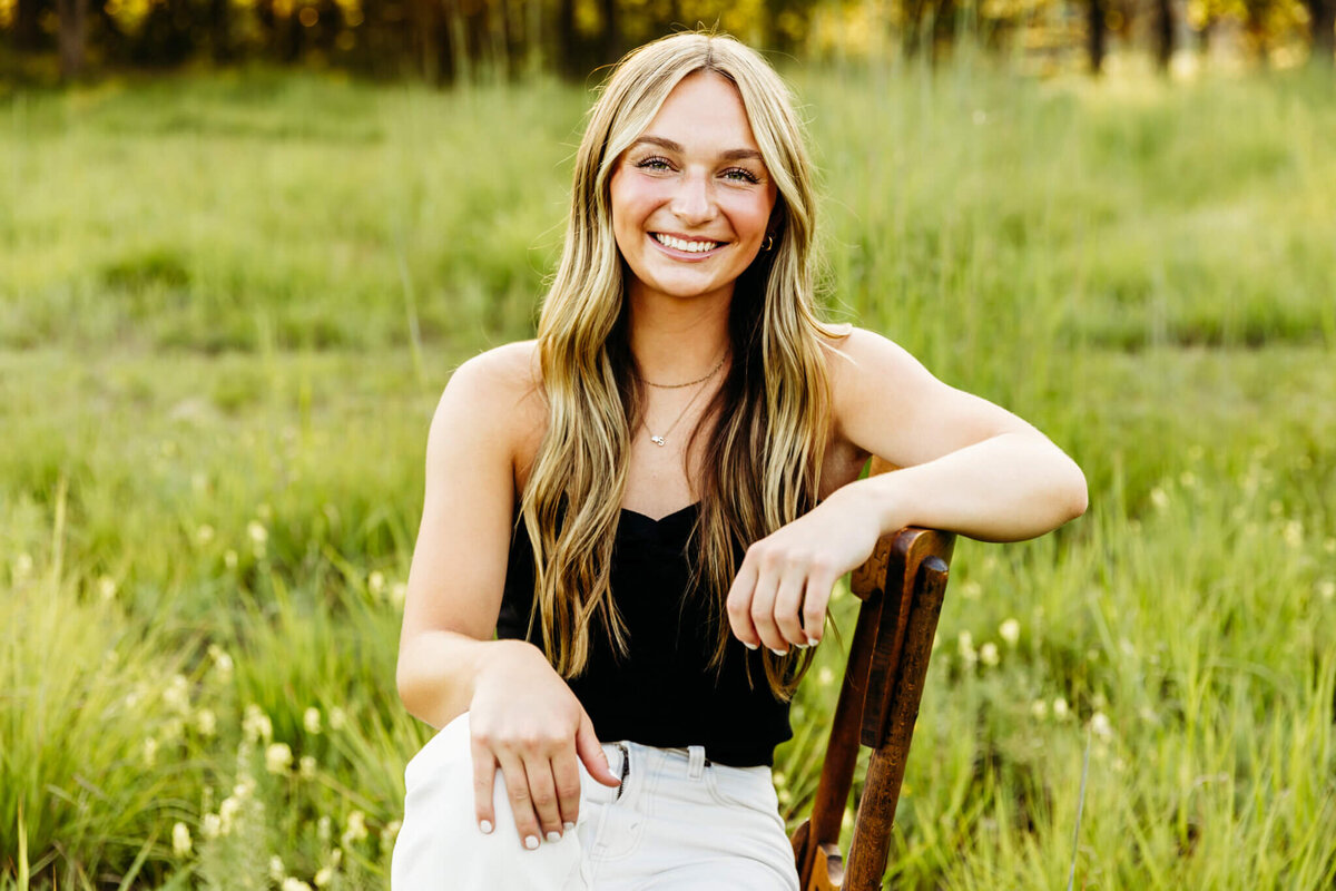 pretty high school senior girl sitting sideways on a wooden chair with arm on the back of chair and legs crossed in a beautiful glowing field near Appleton WI