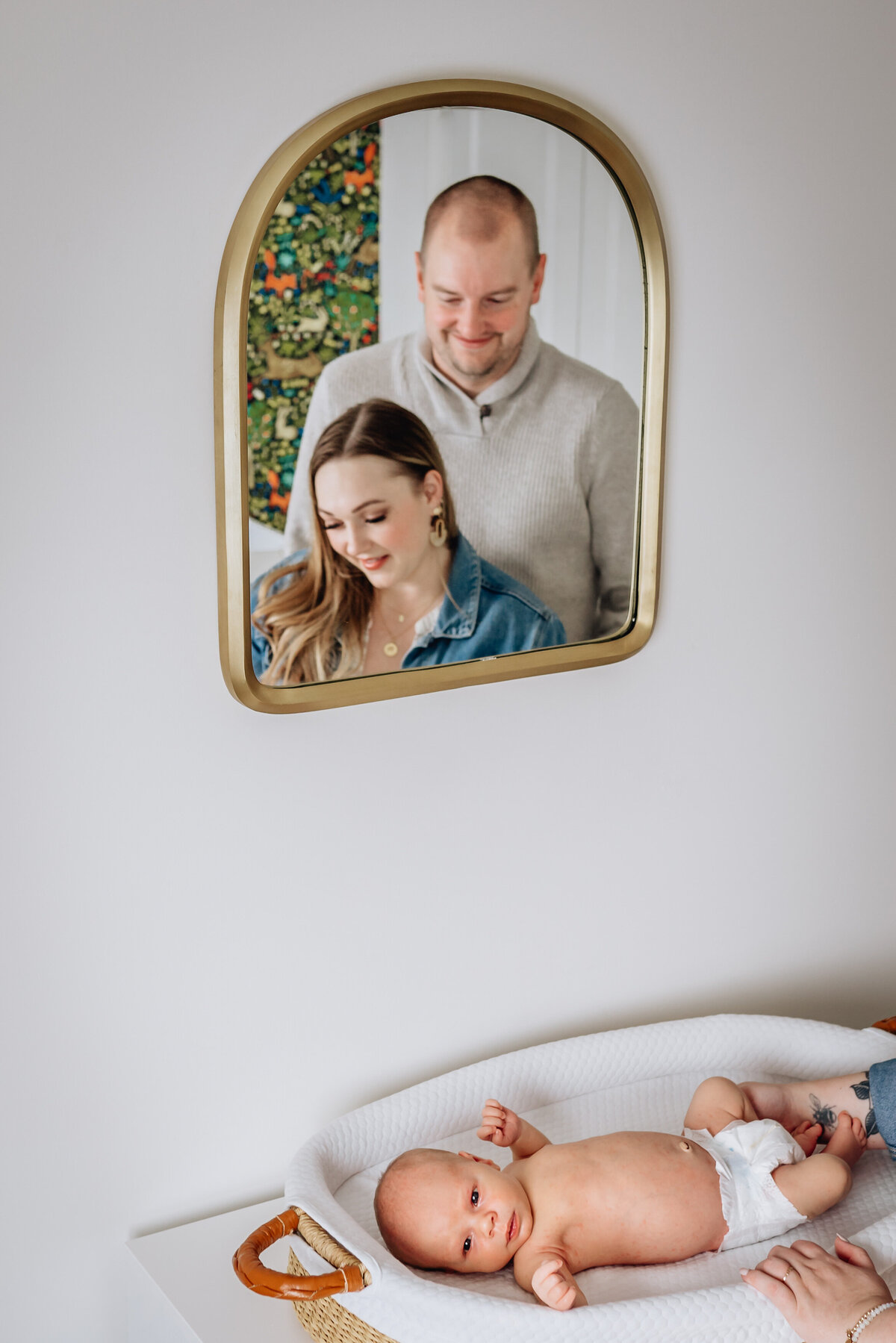The reflection of a smiling couple is seen in a gold-framed mirror as they stand over their baby, who is lying in a basket on a changing table. The baby, wearing only a diaper, gazes upward.