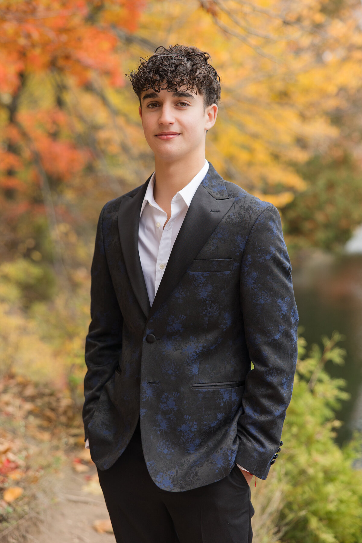 Senior guy in a suit standing in front of beautiful fall colors smiling at the camera