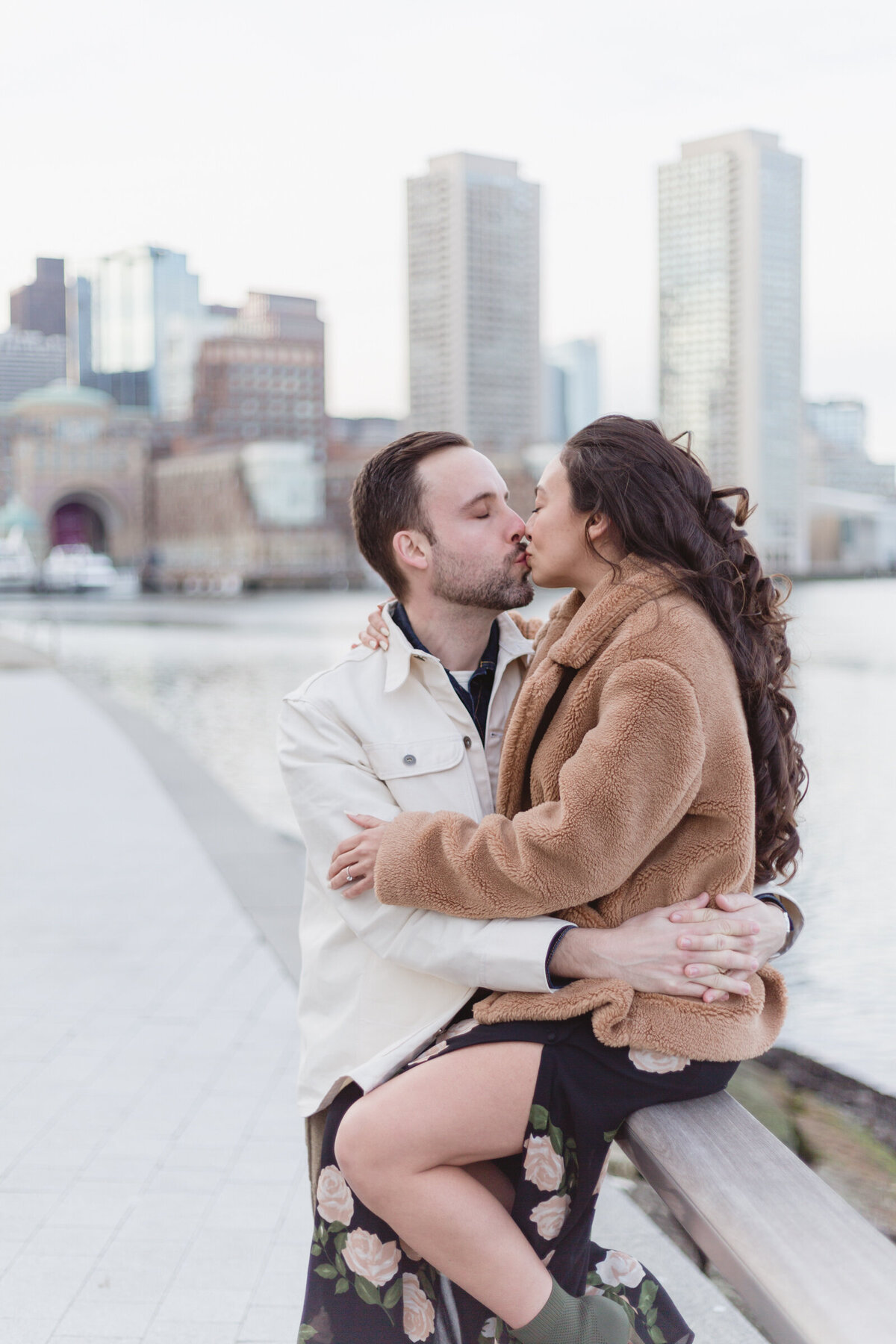Fashionable couple share a kiss in front of the Boston skyline during their engagement session