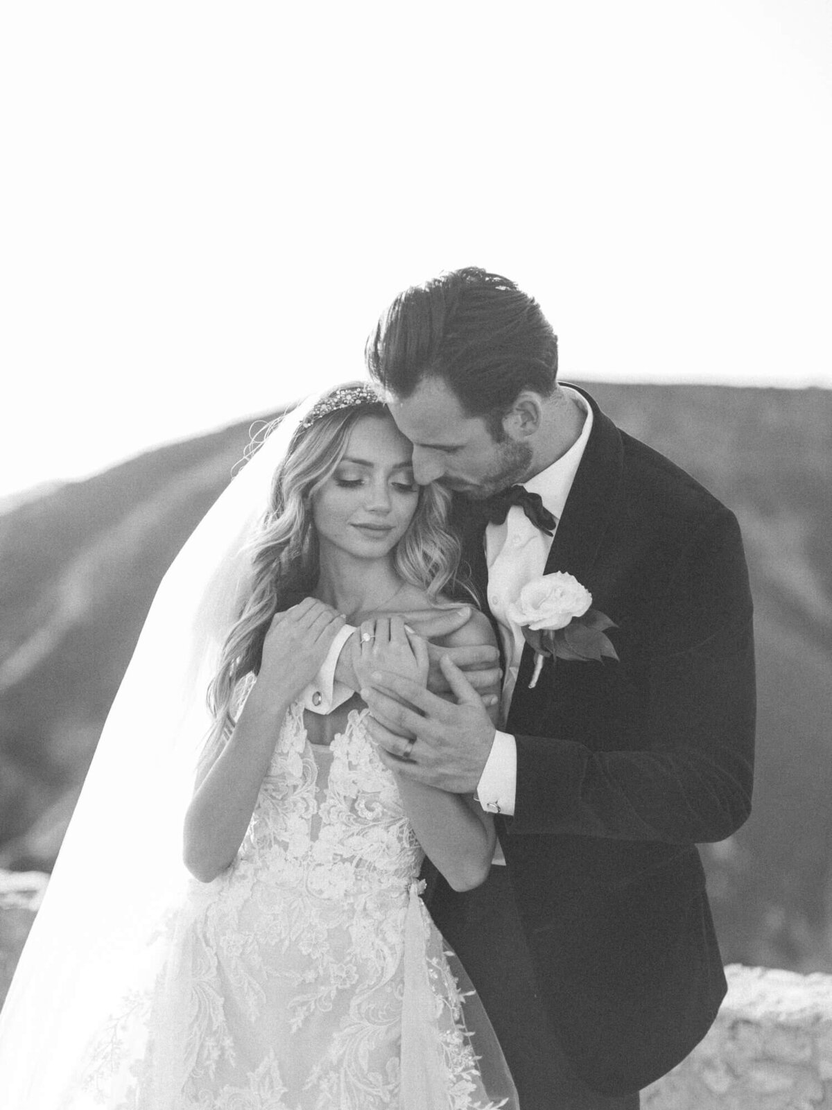 A bride and groom embrace tenderly in an outdoor setting, with soft light illuminating them.