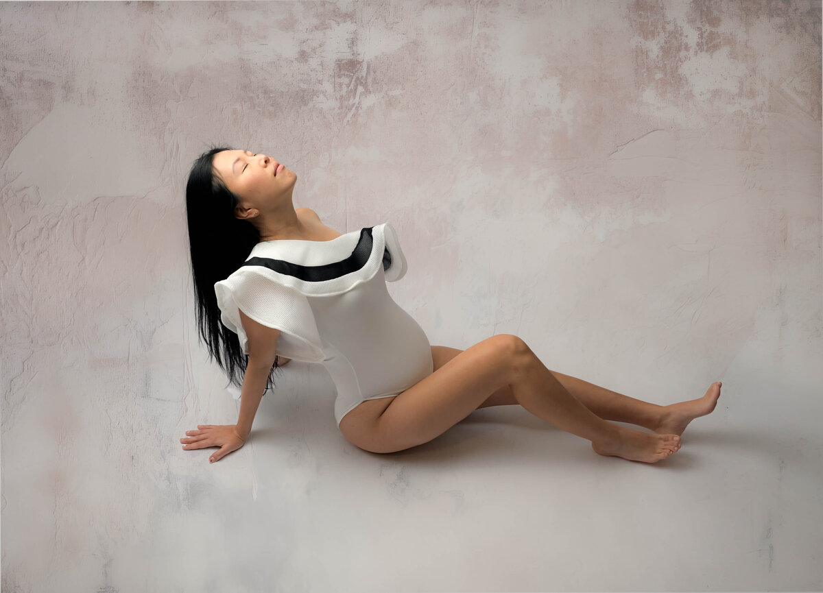 Expecting mother in this studio maternity session sits on the floor in an elegant white bodysuit with legs stretched out, leaning back slightly, and tilts her head as if she's looking to the sky. Image captured by Beth Benecki.