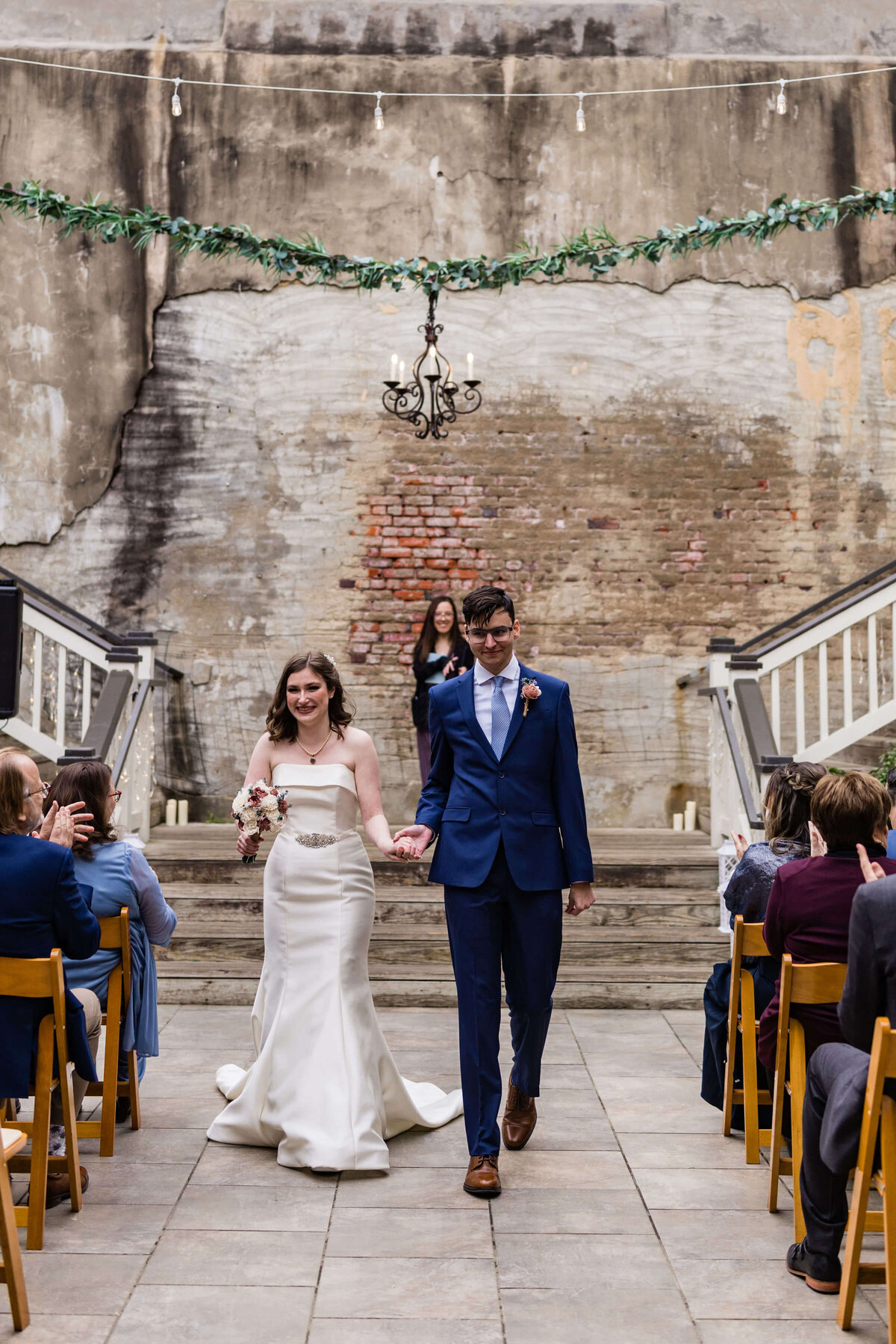 A bride in a white lace gown and a groom in a navy suit walk hand in hand down steps, smiling, after their wedding ceremony. A flower girl and officiant stand in the background near floral decorations.