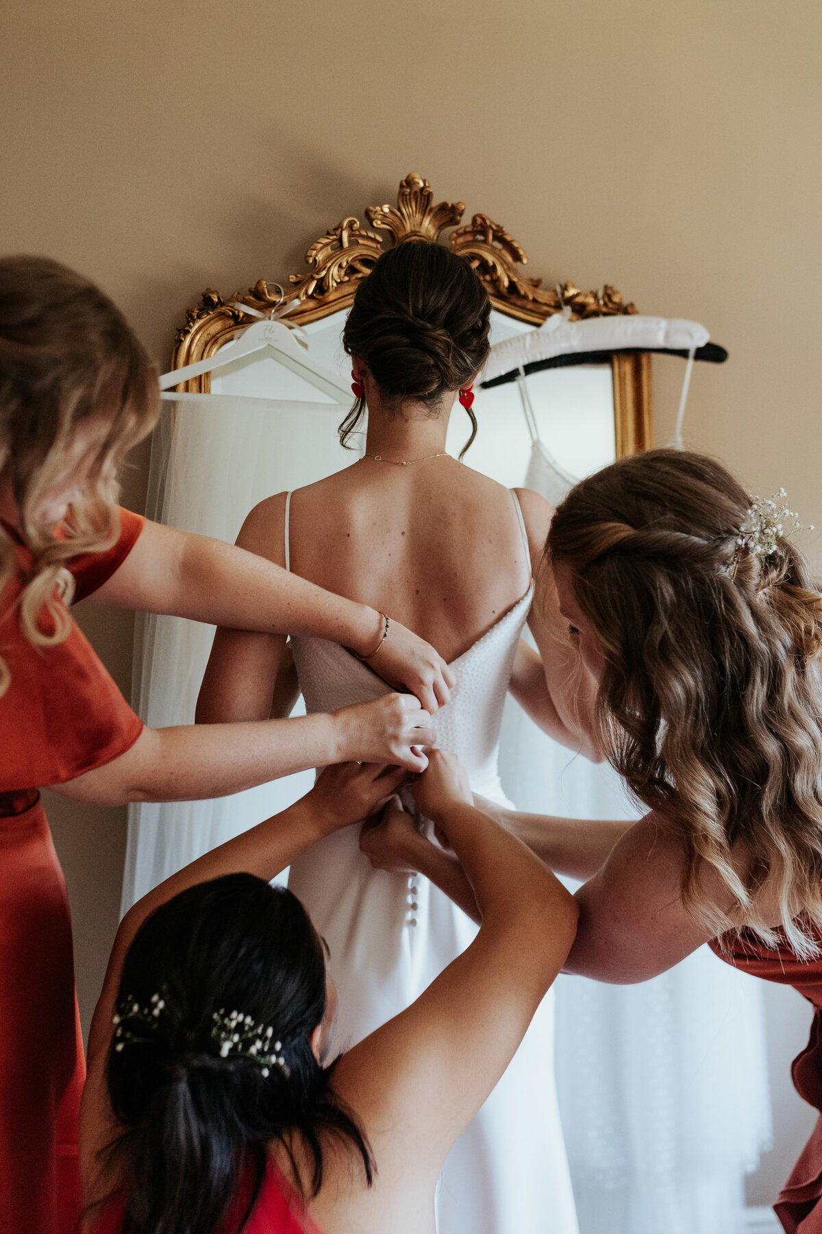 Bride has her dress buttoned up by three bridesmaids infront of a gilded gold mirror