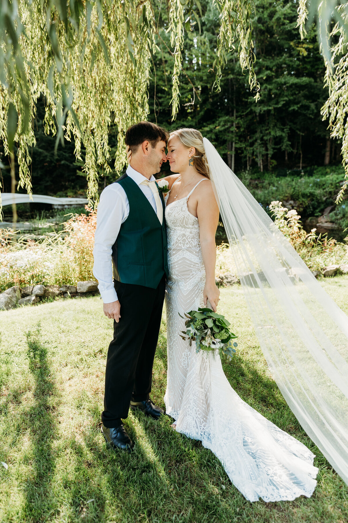 A bride and groom stand under a willow tree touching foreheads